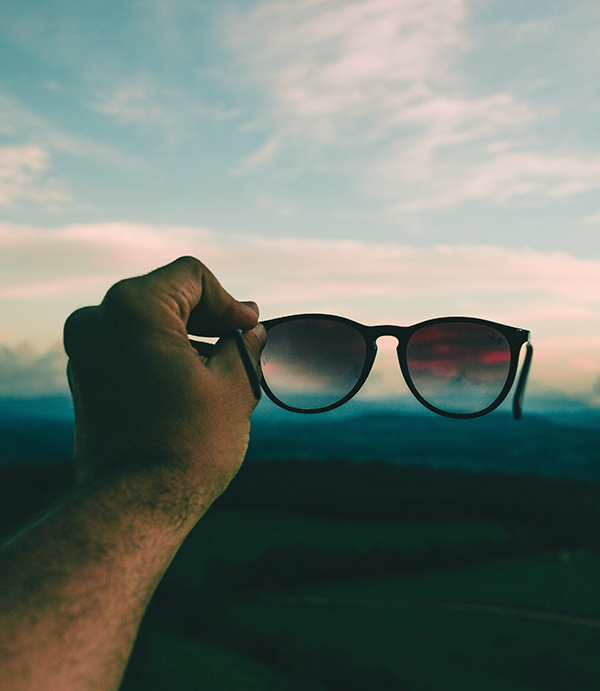 Hand holding out sunglasses with sunset sky in background.