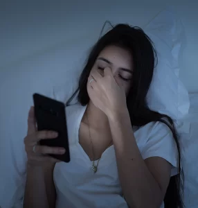 A woman holding her phone and laying on her bed massages the bridge of her nose because of digital eye strain.